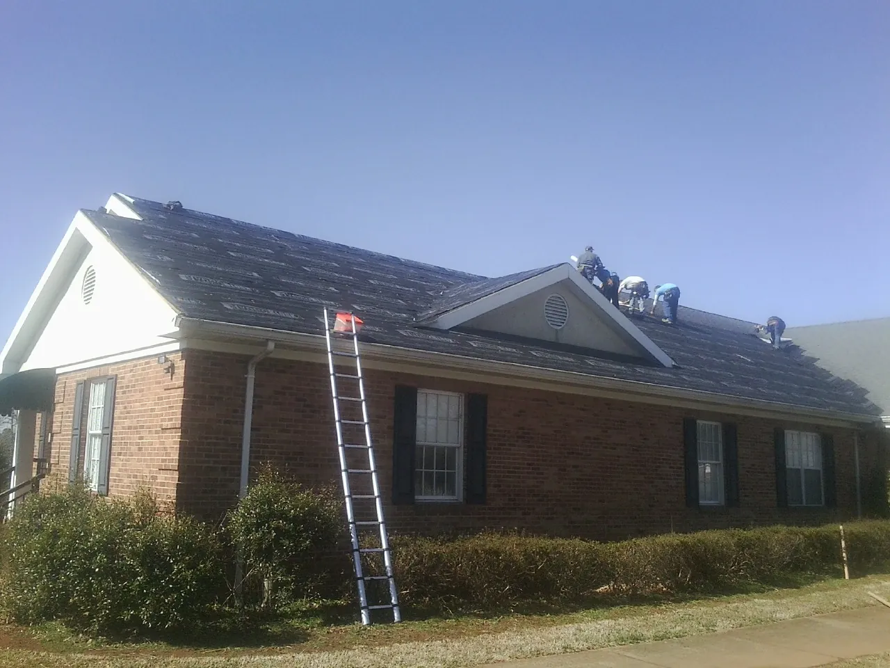 Crew tearing shingles off a brick ranch roof with a ladder set — Metal Roof Repair work by our crew in Harlem