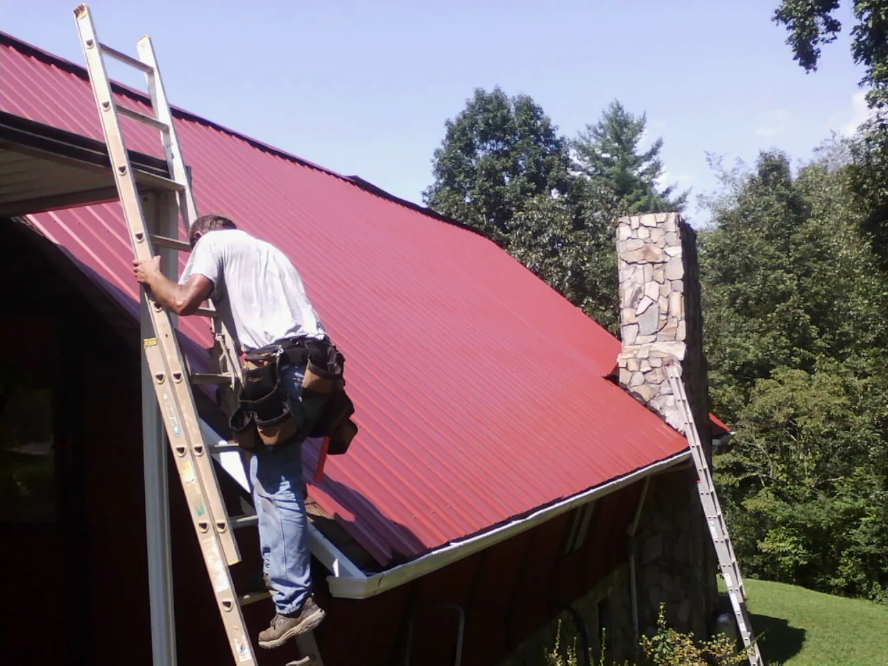 Roofer on a ladder beside a red metal roof and stone chimney — Soffit & Fascia Repair by Roofing Brothers in Harlem